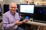 Paul Keeney sits in front of a 3D printer holding two plastic structures