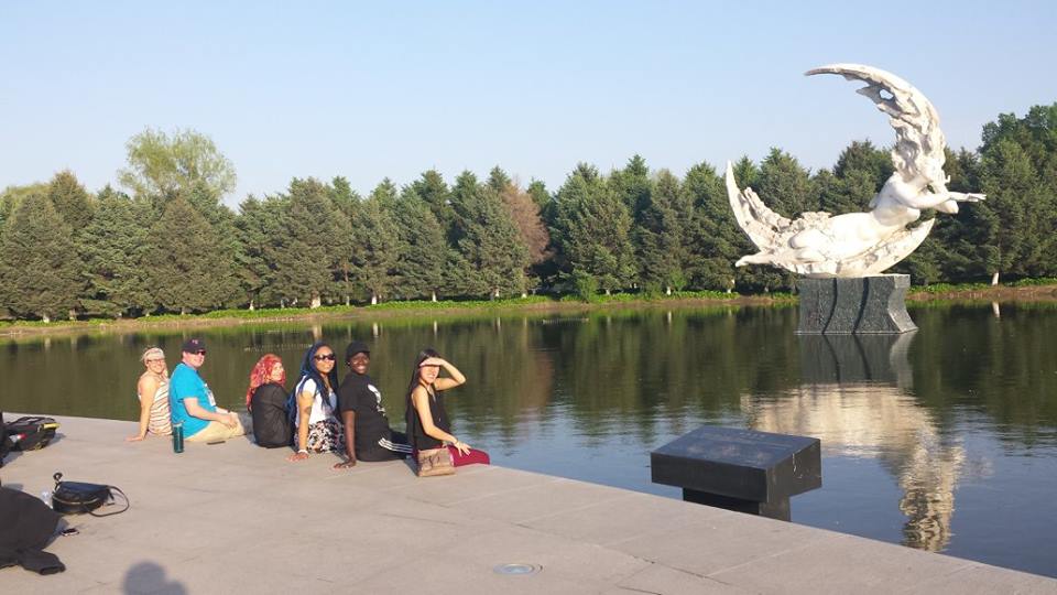 Students sit at the edge of a lake with a sculpture in the background