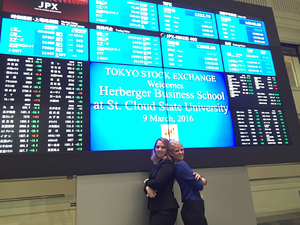 Two students stand in front of a large screen at the Tokyo Stock Exchange welcoming St. Cloud State business students 