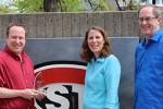 Three people stand with an award in front of the St. Cloud State sign