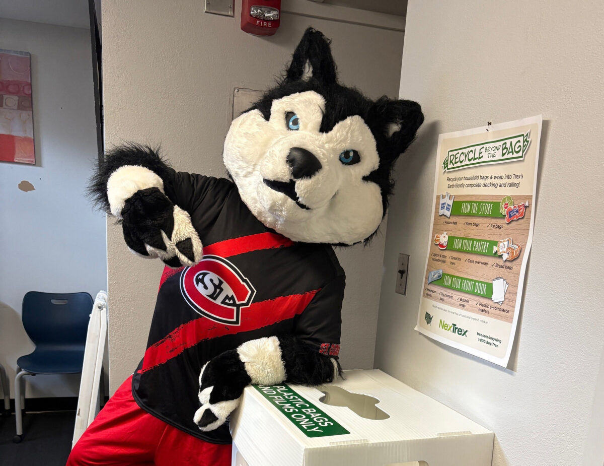 SCSU's mascot blizzard, wearing a red and black striped shirt, points to a white recycle box