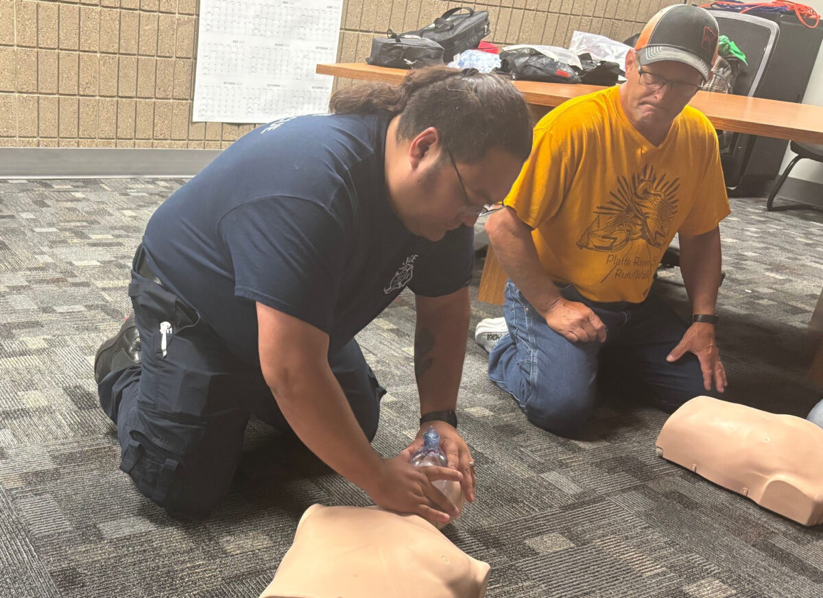A man in a blue shirt and glasses works on a CPR mannequin while a man in a yellow shirt, glasses and hat watches in the background