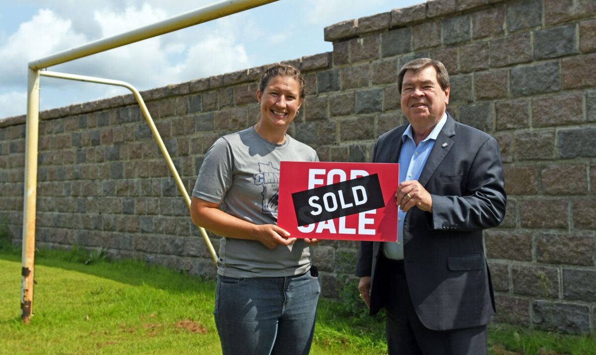 A woman in a grey shirt and jeans and a man in a blzaer and blue dress shirt hold a hold sign with a soccer goal and large granite wall visible in the background