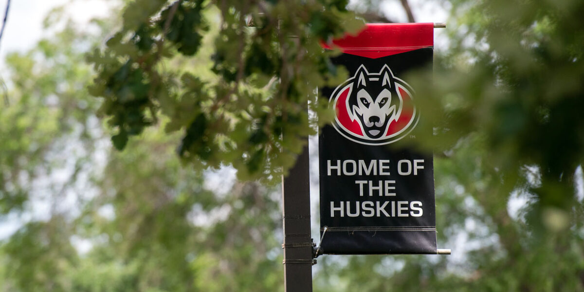 A black home of the huskies banner with the scsu logo is visible through green leaves of a tree