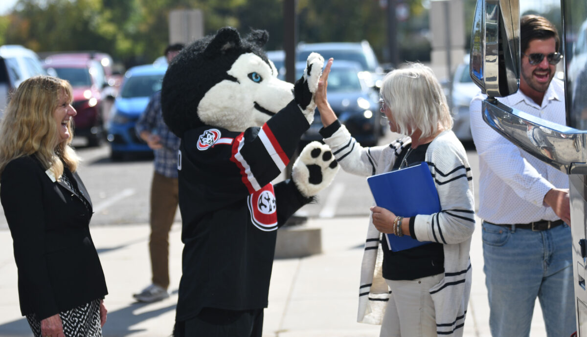 SCSU's mascot Blizzard high fives a woman holding a blue binder as she steps off of a coach bus into the Herb Brooks National Hockey Center parking lot