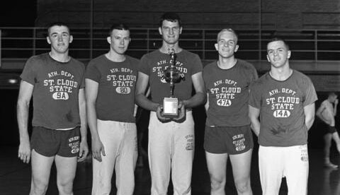 Five men wearing shirts that say Ath. Dept. St. Cloud State smile for a photo while Issy Schmiesing holds a trophy in the center