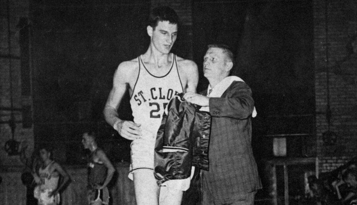 Black and white photo of Issy Schmiesing as he wears a number 25 St. Cloud jersey and is handed a warm up jacket by coach Red Severson with players visible in the background