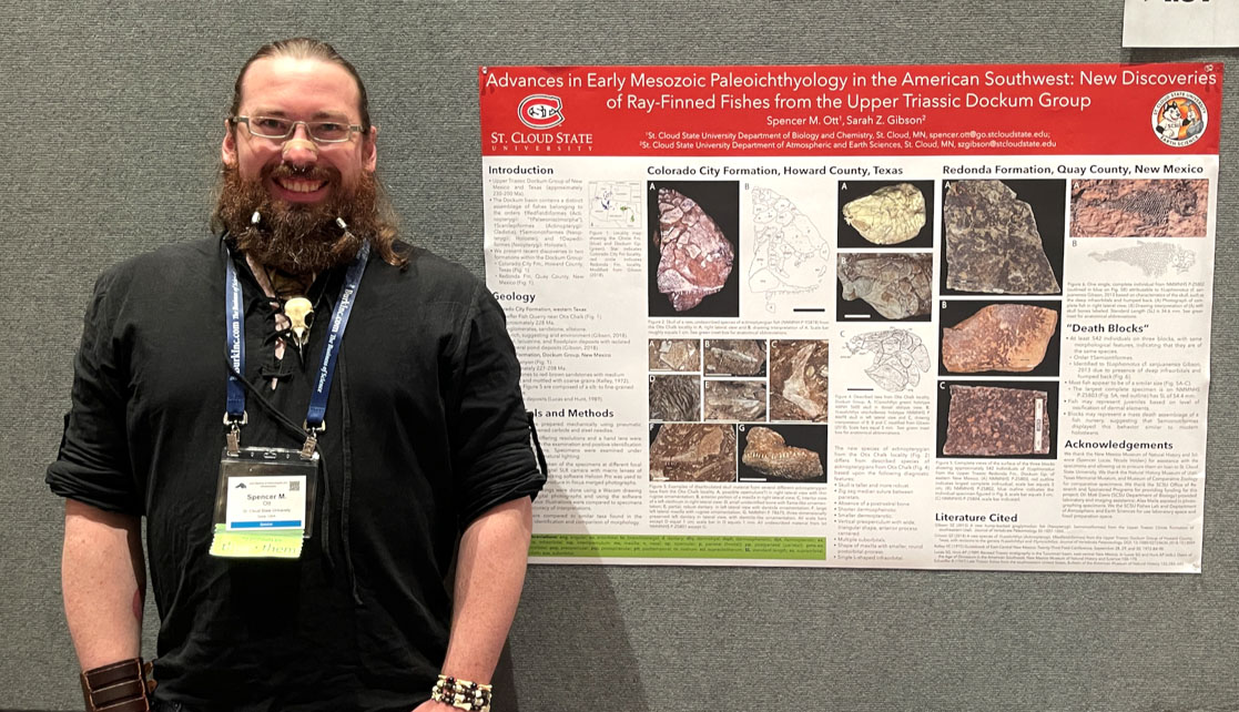 A man with a black shirt, glasses and beard smiles at the camera next to a poster with various pictures of fish fossils
