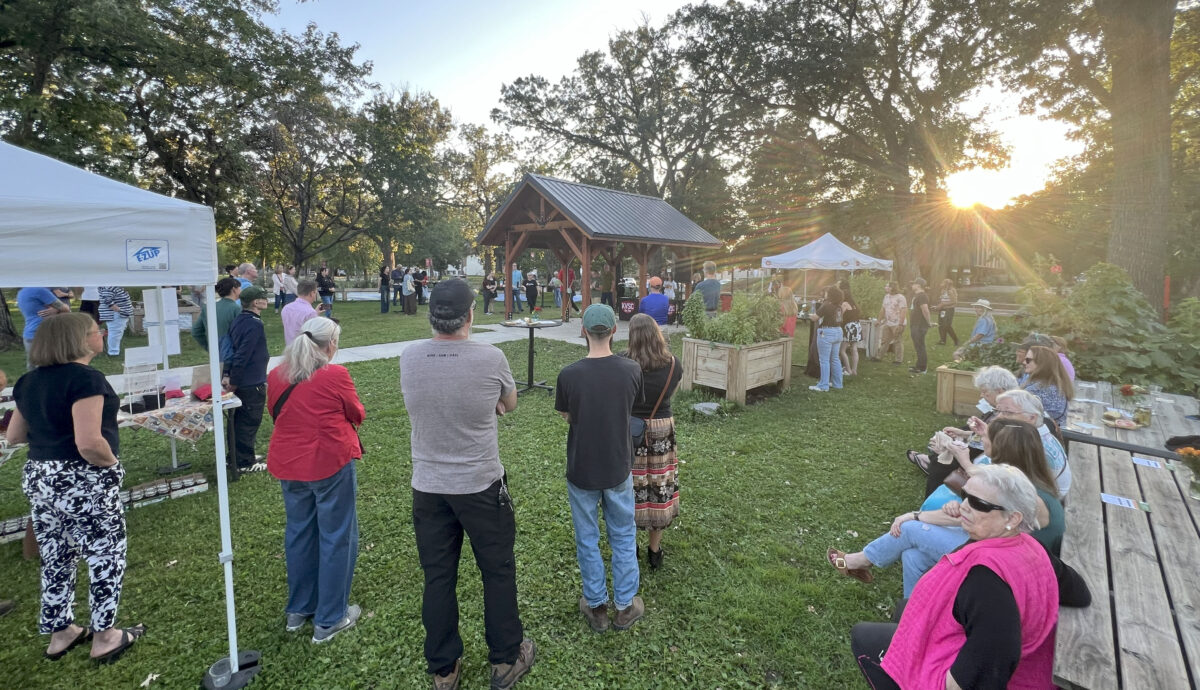 A crowd of people face the Community Garden pavilion while the sun sets.
