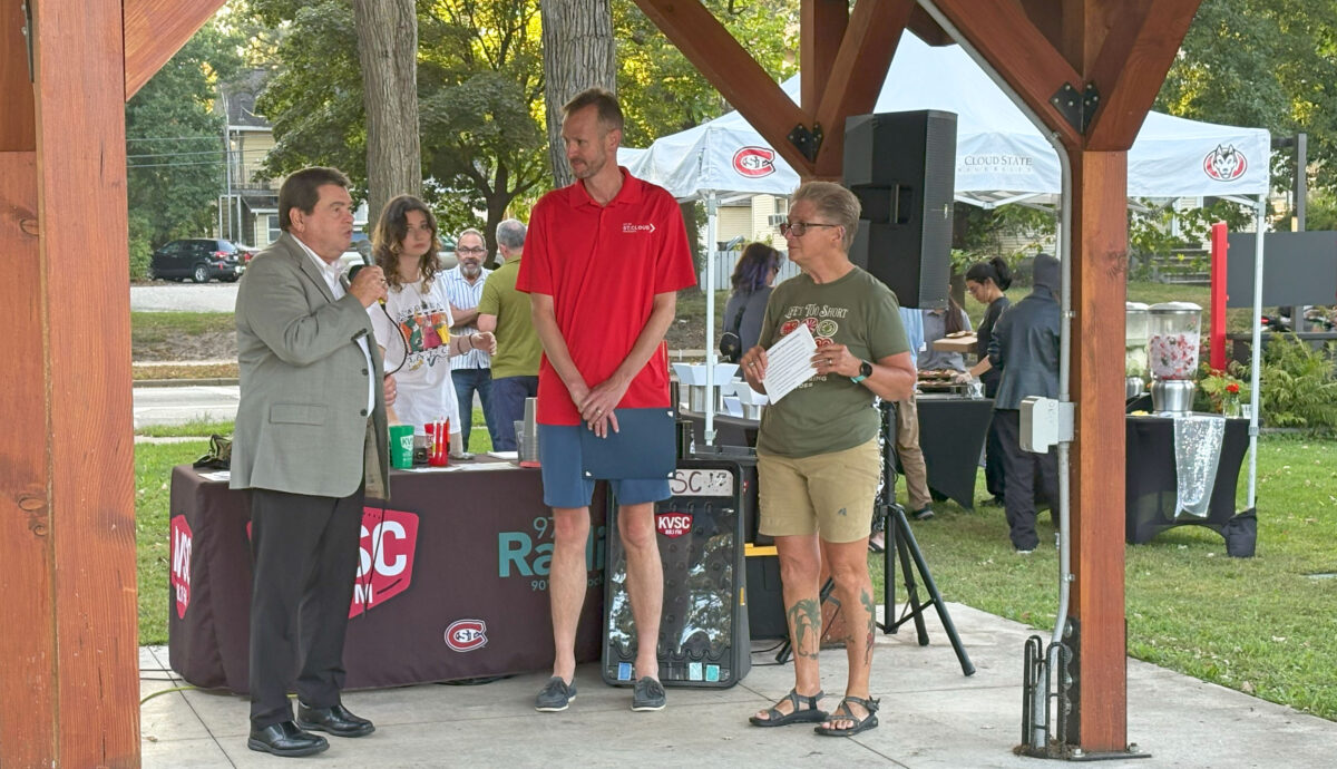 A man in a suit speaks into the microphone while a man in a red shirt and a woman in a green shirt and khaki shorts stand under a wood shleter with SCSU tent visible in the background