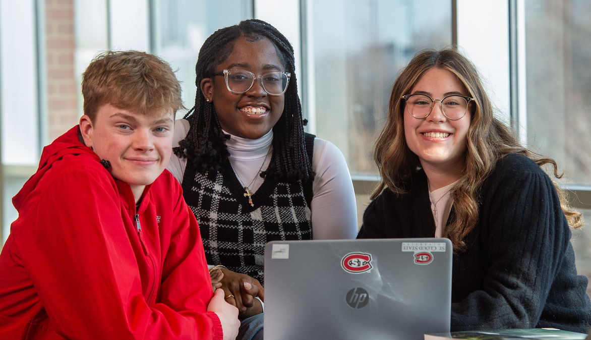 A man in a red sweatshirt, a woman with black hair, glasses, and a sweater and a woman with brown hair, glasses and sweater smile at the camera with a laptop with SCSU logos in front of them
