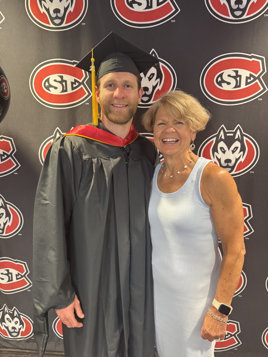 Clark Kuster smiles wearing a black graduation cap and gown next to his mom Debbie wearing a dress while standing in front of a black background with SCSU logos