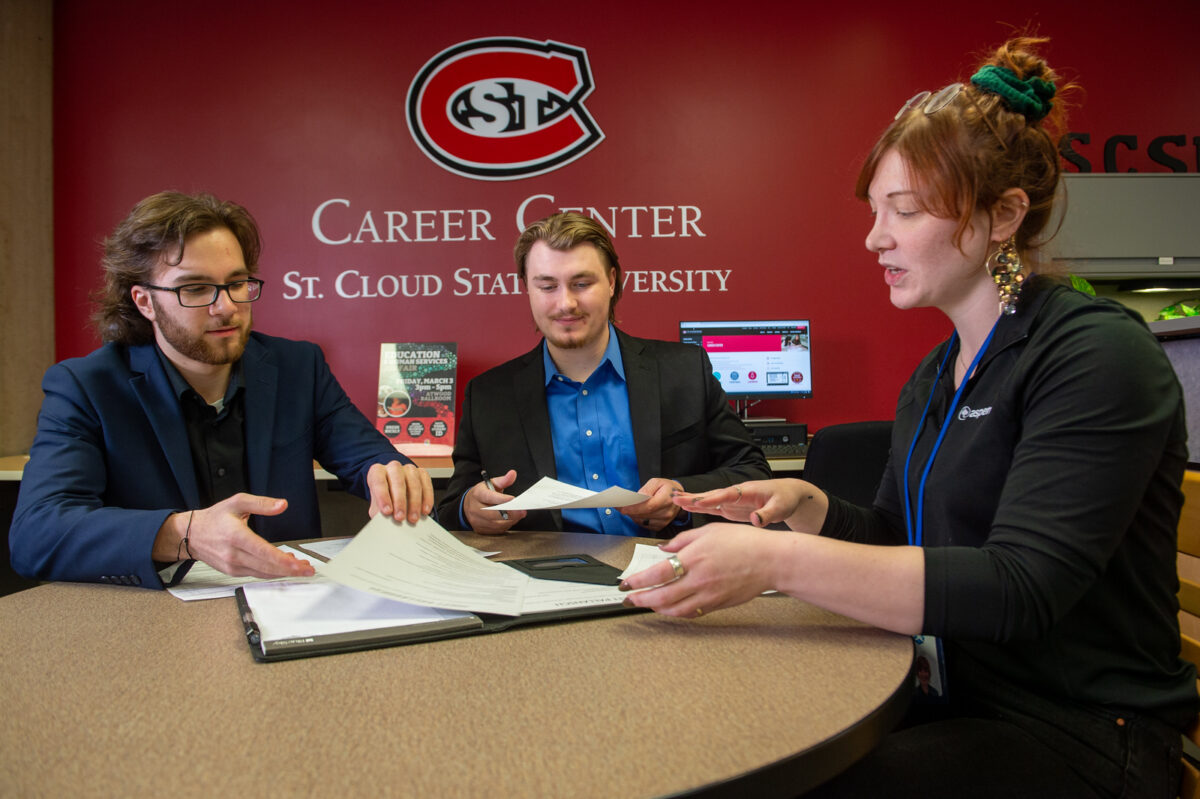 Two men in suits look at pieces of paper while a woman in a black shirt and a bun speaks