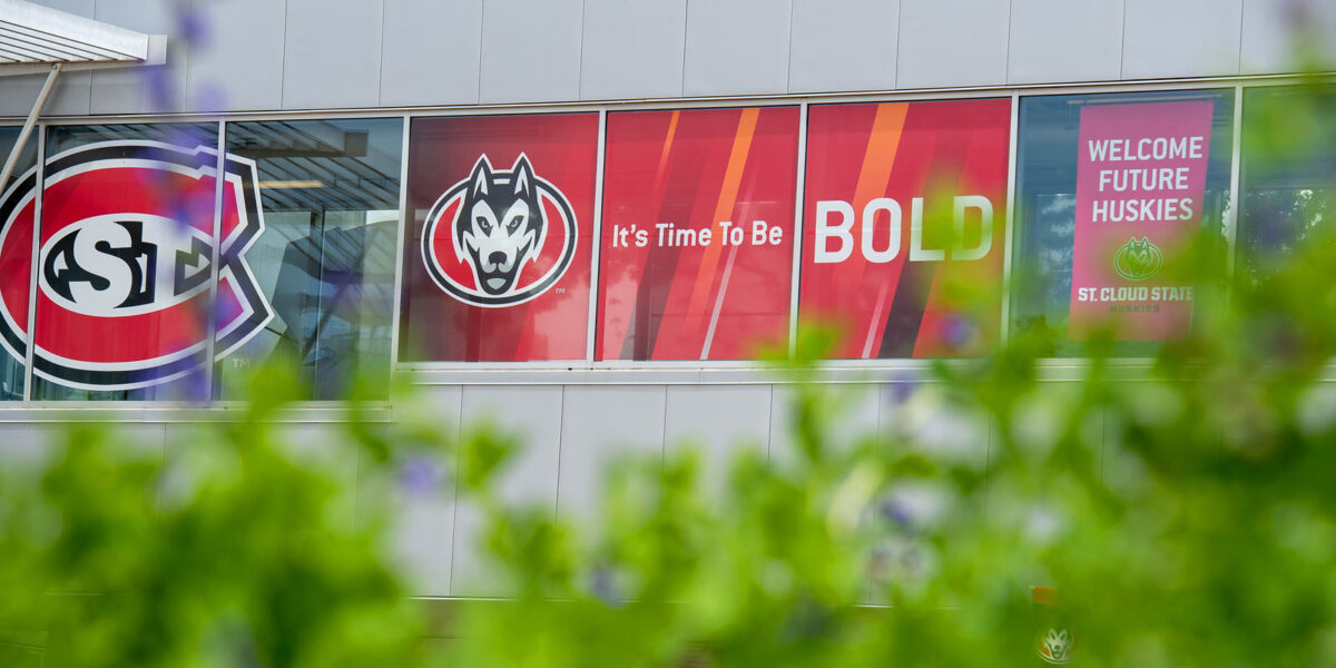 The Atwood Memorial Center skyway includes a St. C logo, Husky Head logo with It's Time to Be Bold Graphic and a Welcome Future Huskies banner with a green bush out of focus