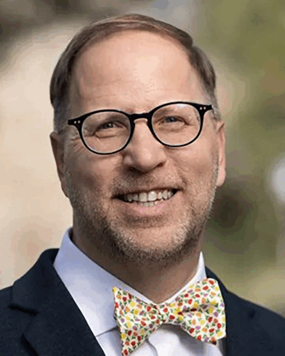 A man with glasses, a beard and a polka dot bowtie wears a suit while smiling for a headshot