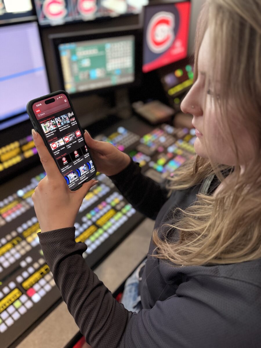 A woman with blonde hair holds a phone showing the UTVS app within the studio's control room