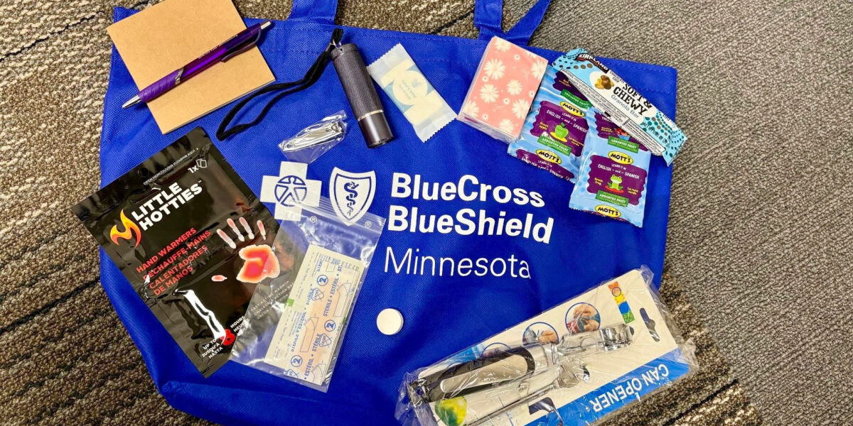 A BlueCross BlueShield Minnesota blue bag has various items like food, a can opener, and hand warmers sitting on top of it