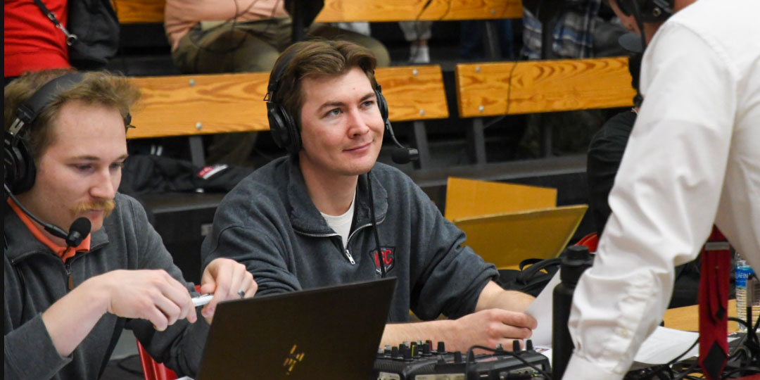 Two men sit at a table with headsets on while they interview a coach in a white dress shirt