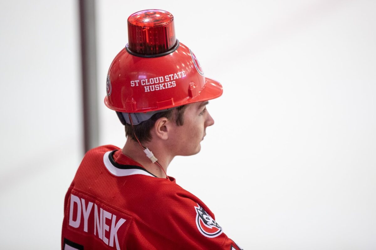 Carl Goenner wears a red SCSU hockey jersey with the last name Dynek on it while wearing a St. Cloud State Huskies red hat with a light on top
