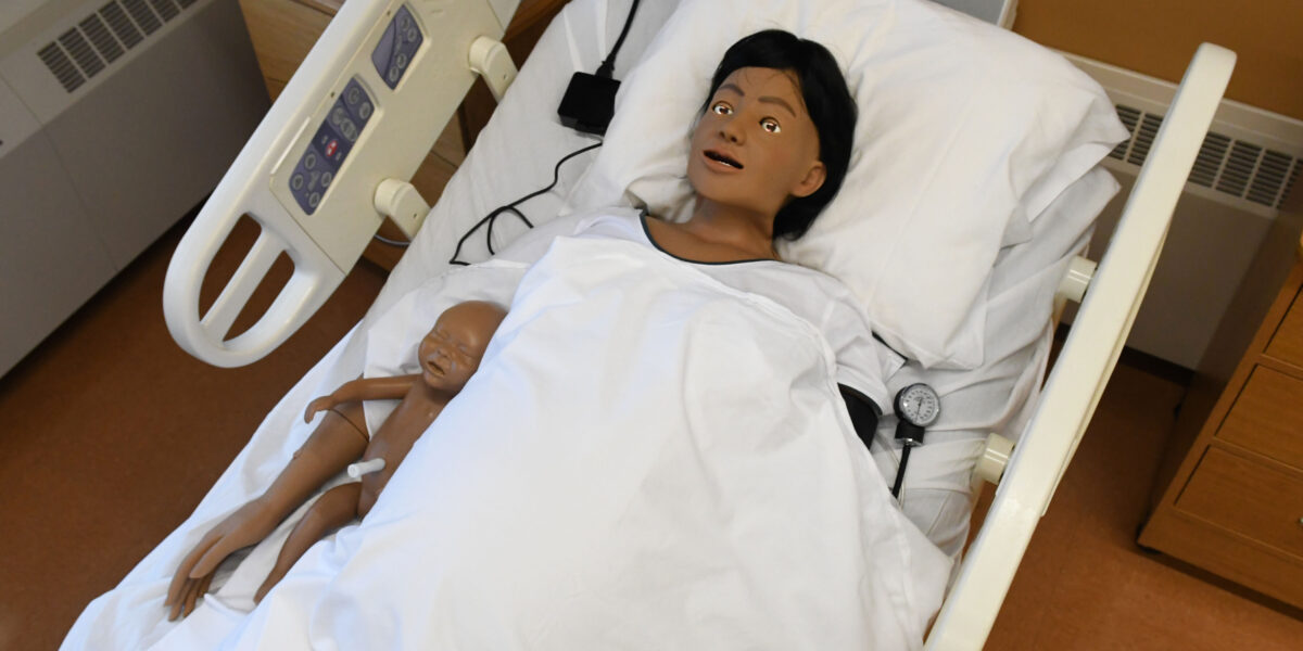 A manikin with dark hair holds a baby while laying in a hospital bed