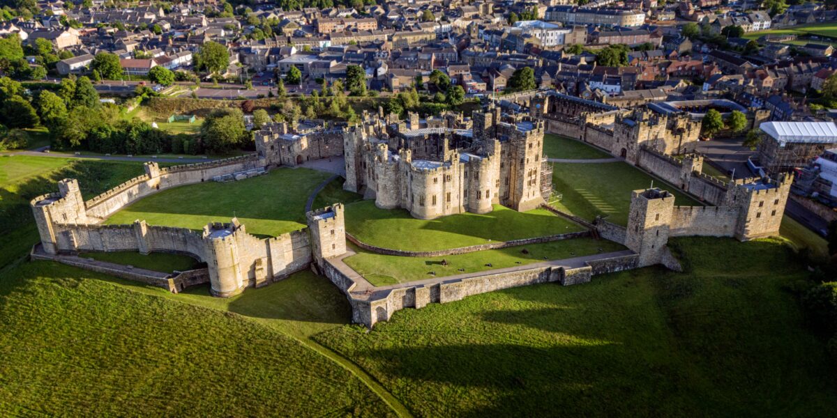 An aerial photo of Alnwick Castle with its large outer wall and green lawns. The town of Alnwick is visible nearby.
