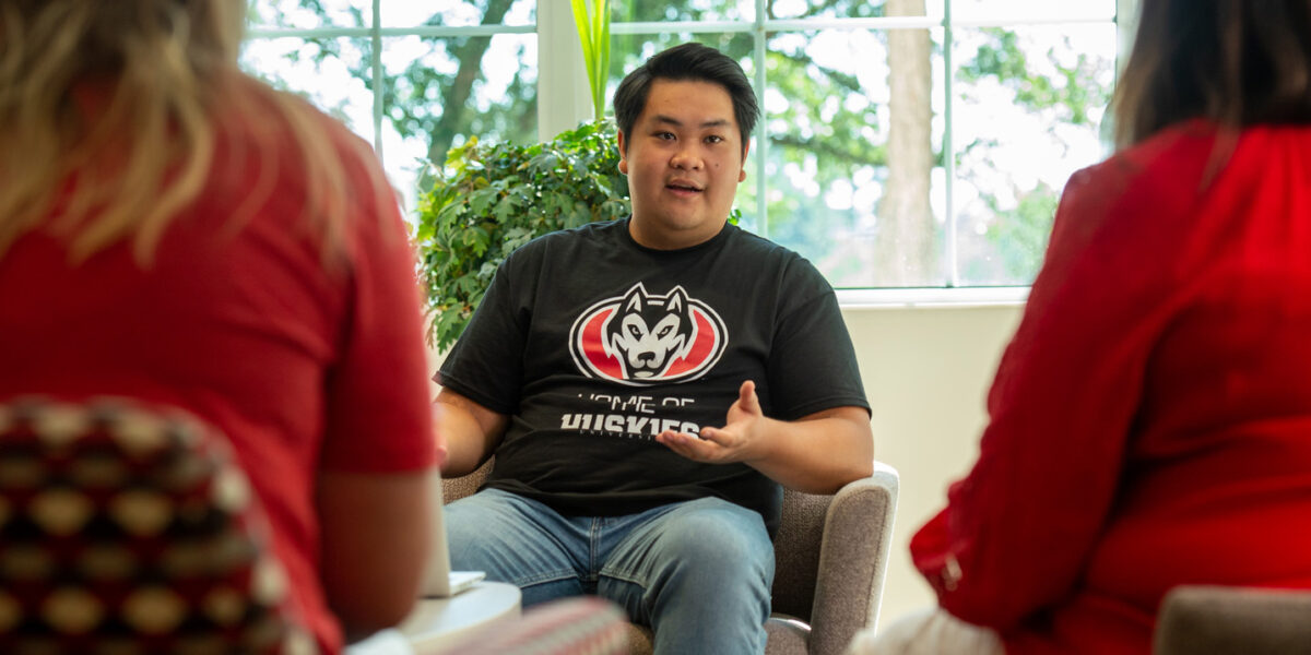 A man in a Home of the Huskies black shirt and blue jeans sits in a chair with a window behind him and two women in red shirts out of focus