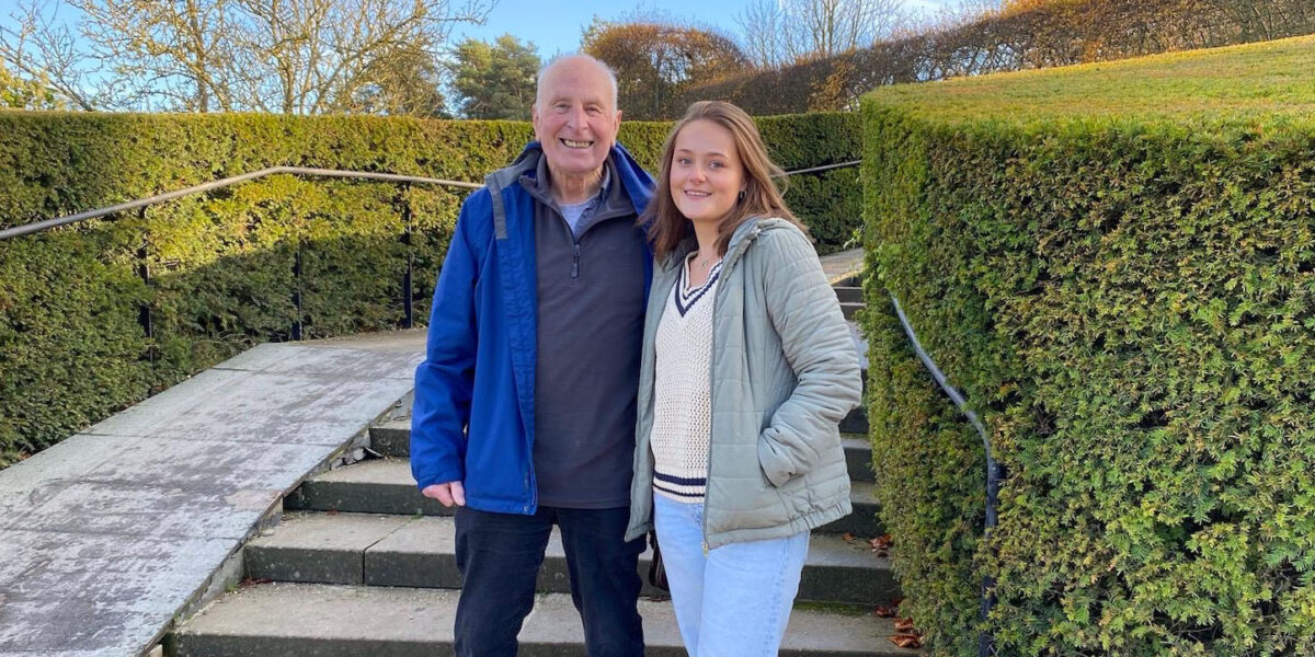 Terry Smith, wearing a blue jacket and a quarter zip, stands next to Katie Tompkins, wearing a jacket and sweater, smile for a photo on a staircase next to tall green hedges