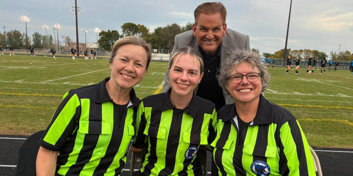 Three women in yellow and black striped shirts smile for a photo in front of a man in a grey blazer with a soccer field in the background