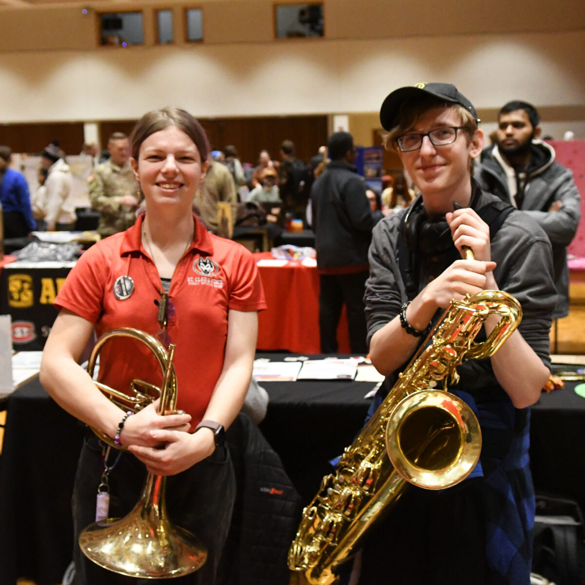 A woman in a red polo holds a horn and a man in glasses and a hat holds a saxophone as they look at the camera