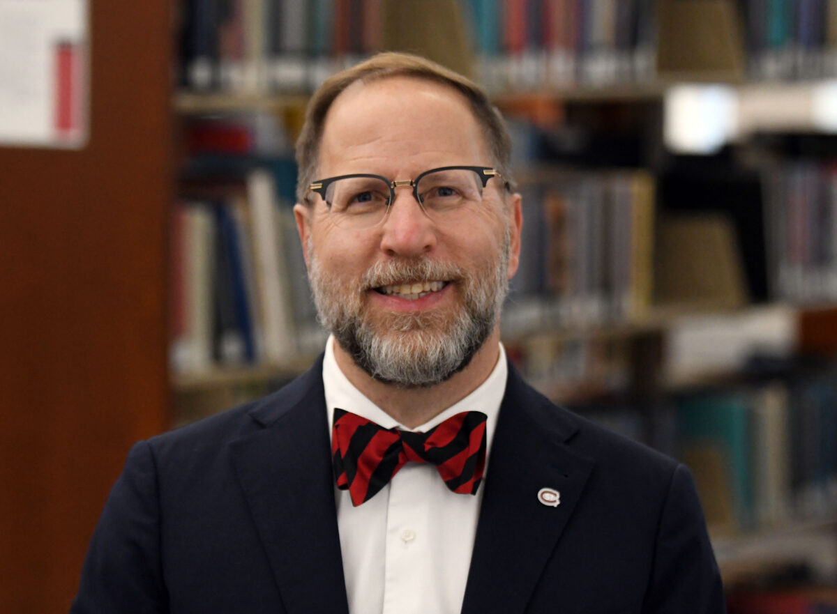 Dr. Gregory Tomso smiles in front of a shelf of books while wearing a suit and red and black bowtie