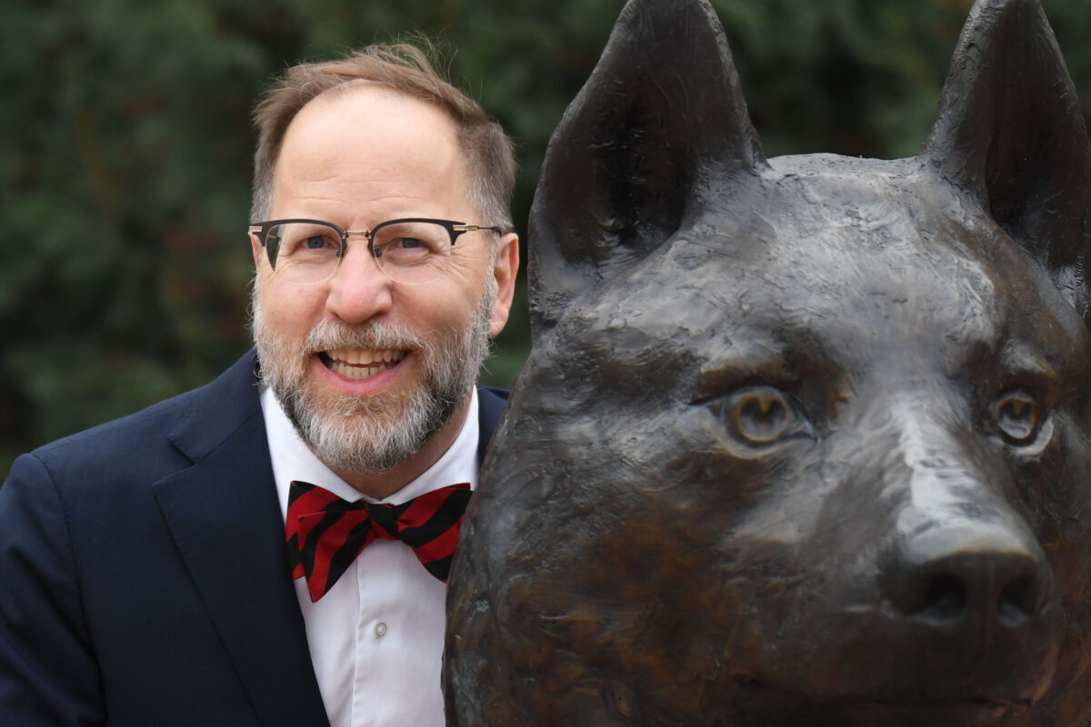 Dr. Gregory Tomso, wearing a bow tie and suit, smiles next to the husky statue in Husky Plaza