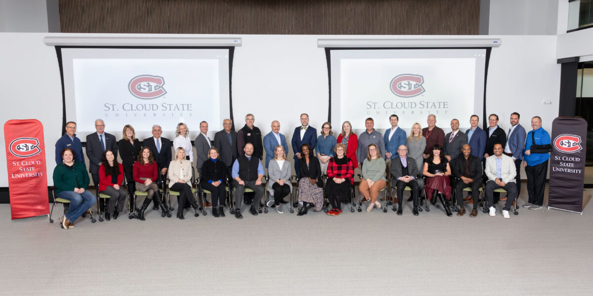 A group of 35 people smile for a photo next to St. Cloud State University banners and screen backgrounds