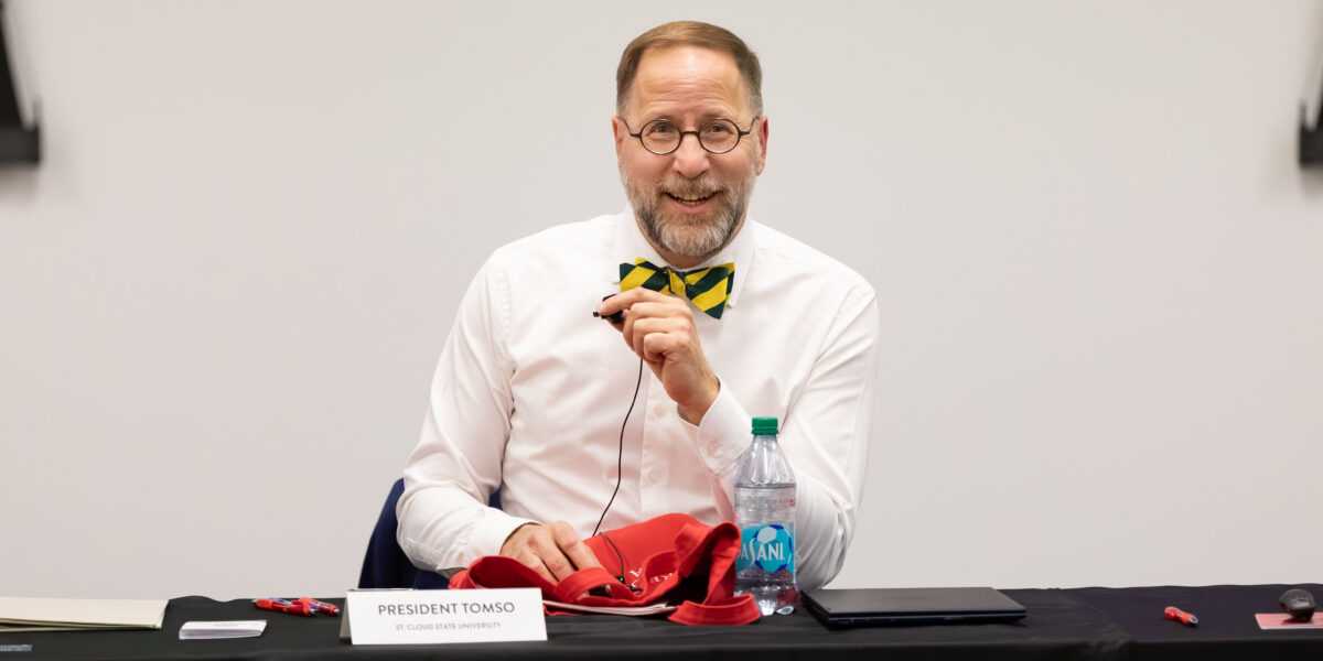 President Tomso, wearing a white shirt and black and yellow bow tie, smiles during a meeting.