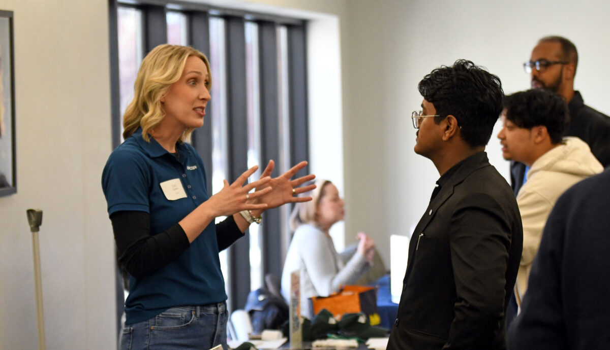 A blonde woman in a blue t shirt and jeans speaks to a students with glasses and a suit at the Winter Career Fair
