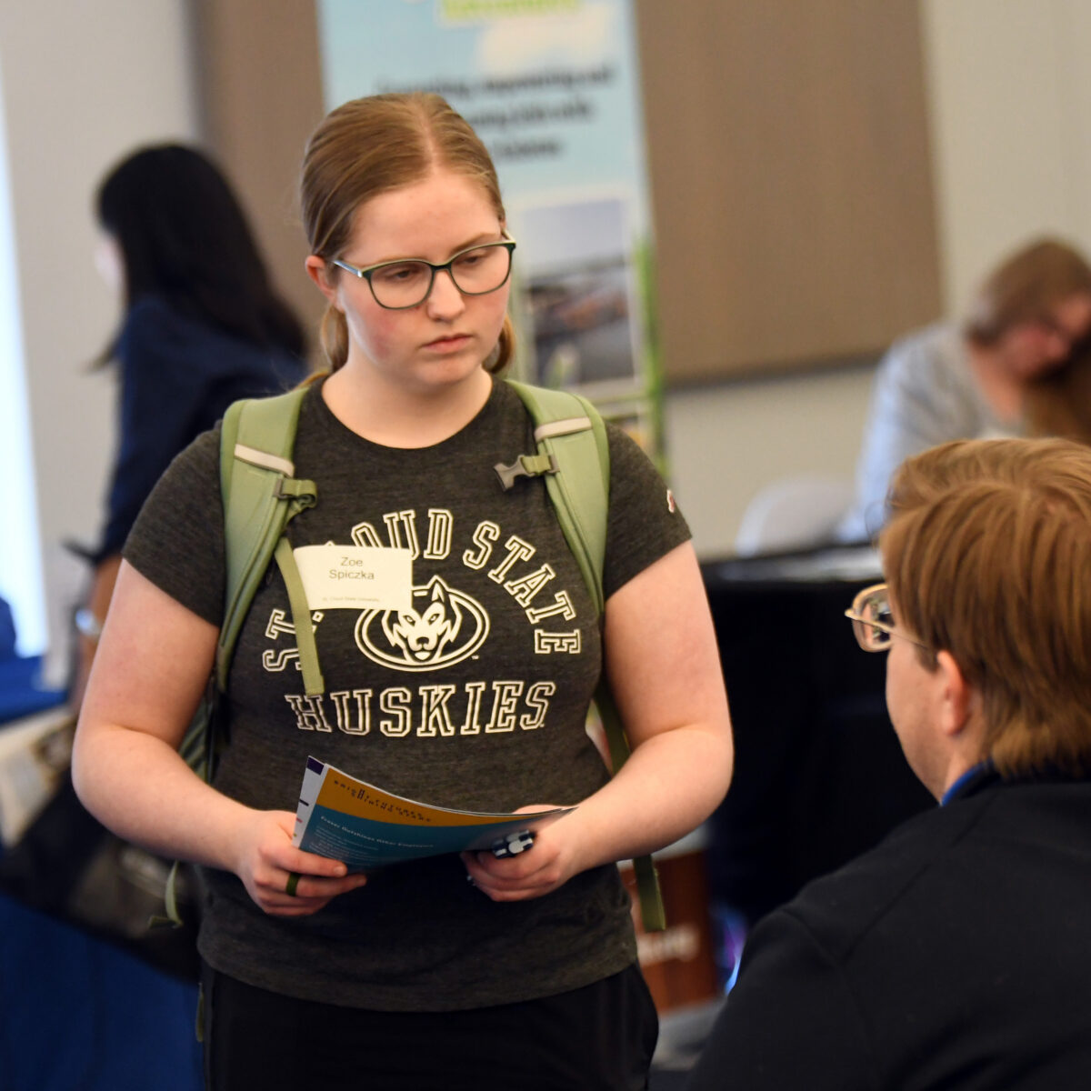 A woman with glasses and a St. Cloud State Huskies shirt speaks to a man in glasses.