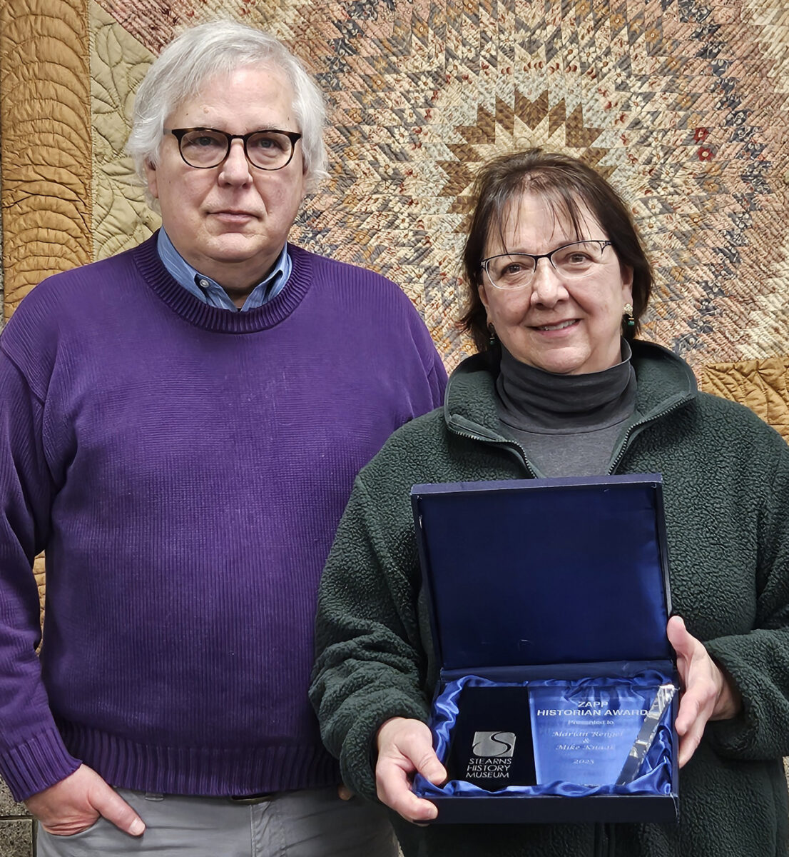 A man with glasses and a sweater stands next to a woman holding a box with the Zapp Historian Award