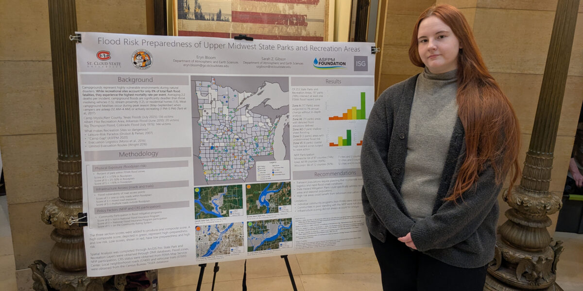 A woman with long red hair and a sweater stands next to a poster with a large map of Minnesota, Iowa and Wisconsin visible