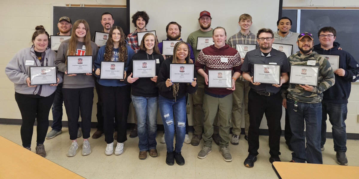 A group of 16 students in two rows in a classroom all smile for a photo while holding certificates