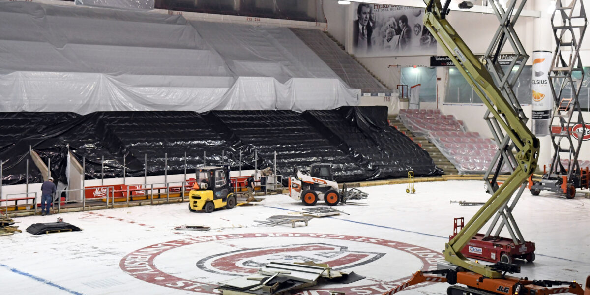 A forklift and skid steer are visible during construction of the Herb Brooks National Hockey Center. Other large equipment is on the ice, while seats are covered with black and white tarps