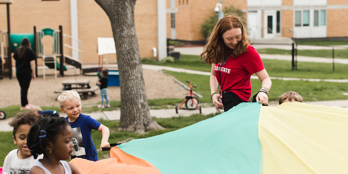A woman with red hair and a red St. Cloud State shirt holds a multi colored parachute with four children present. A playground is visible in the background
