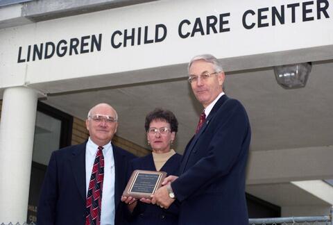 Steve and Jeannie Lindgren, wearing formal attire and glasses, stand next to SCSU president Bruce Grube, wearing a suit and tie. The words Lindgren Child Care Center are visible above their heads.