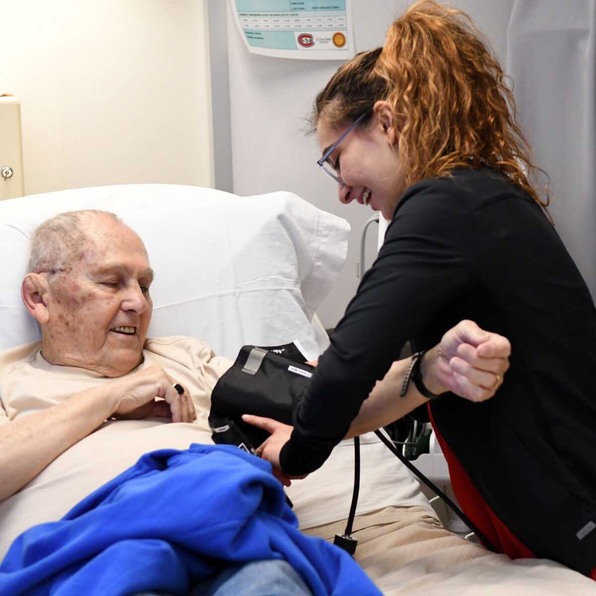 A woman in black scrubs puts a blood pressure monitor on a man sitting in a hospital bed