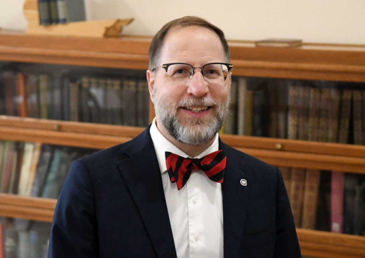 President Tomso, wearing a suit and red and black bow tie, smiles for a photo in front of a shelf of books