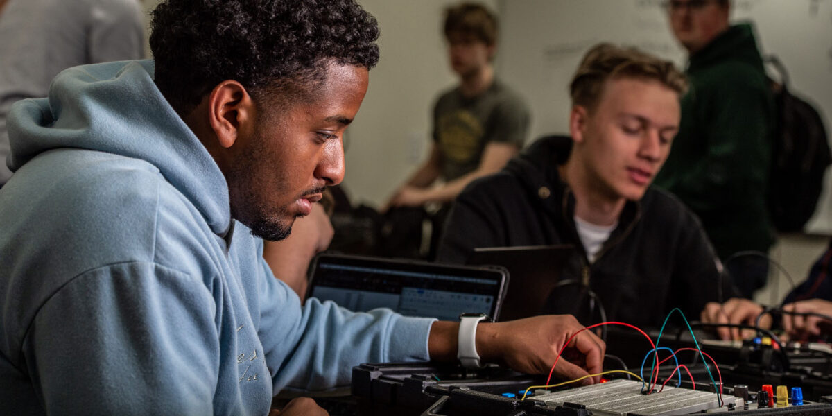 A man in a sweatshirt works on a computer board with other students visible in the background