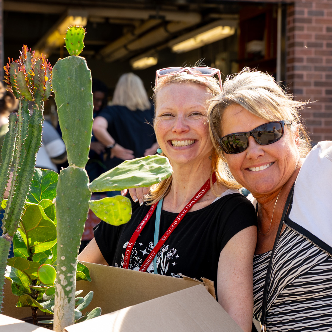 Two women smile at the camera while holding a box of plants