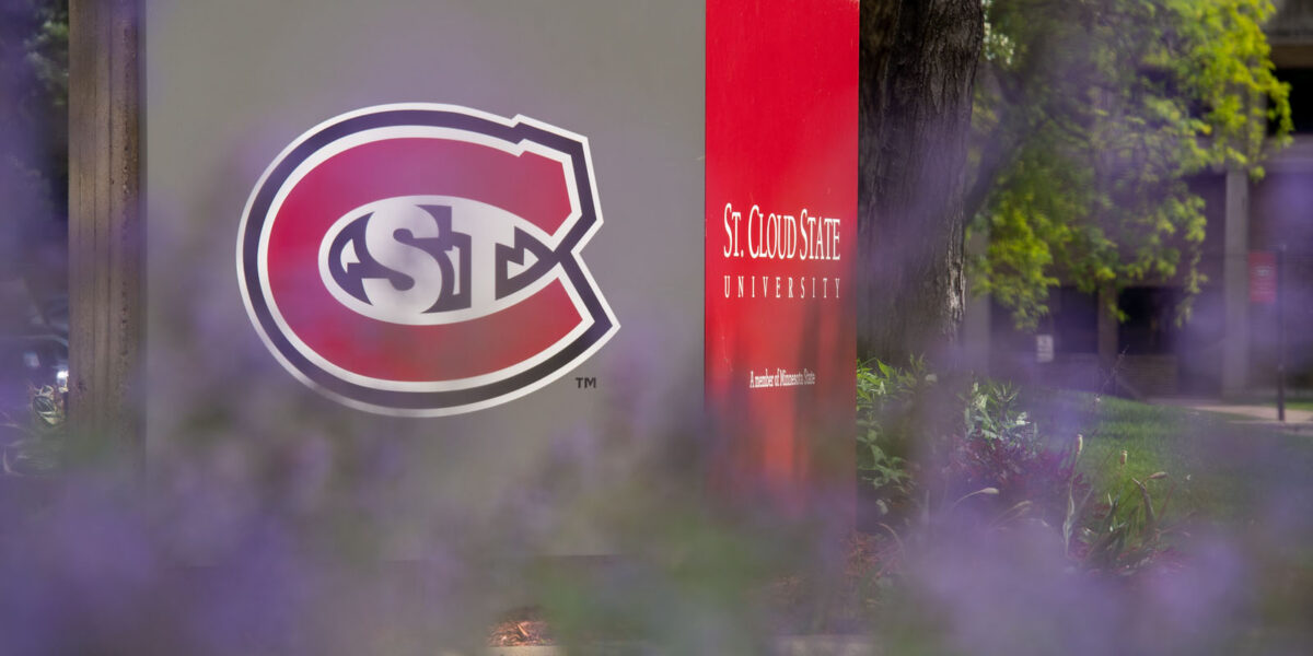 The St. C logo on a St. Cloud State University sign is slightly blurred by purple flowers on a summer day