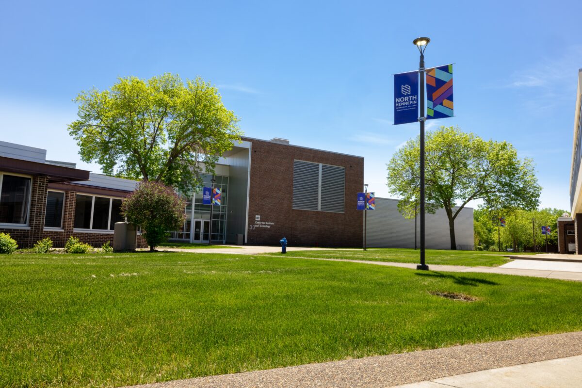 North Hennepin banners are visible on a lightpole next to a grassy area on a sunny day
