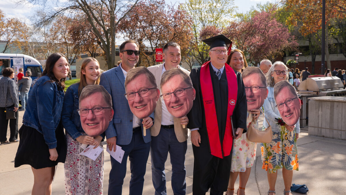David Silker, dressed in a black graduation gown, smiles next to family members with large cutouts of his face