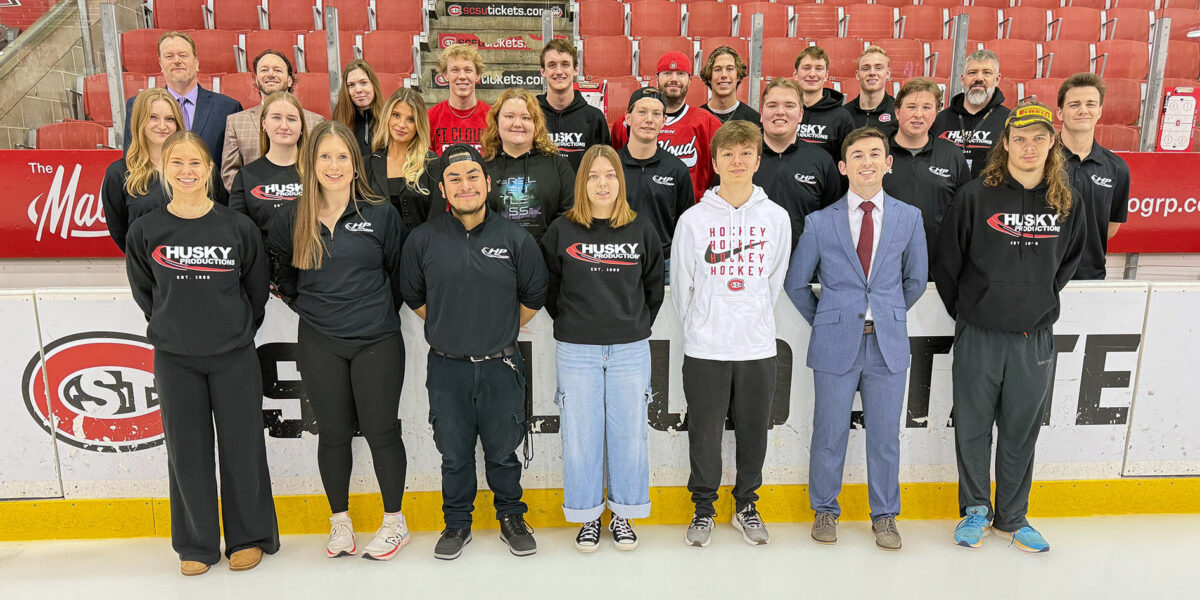 A group of 25 people, many wearing Husky Productions shirts, smile for a photo in the Herb Brooks National Hockey Center