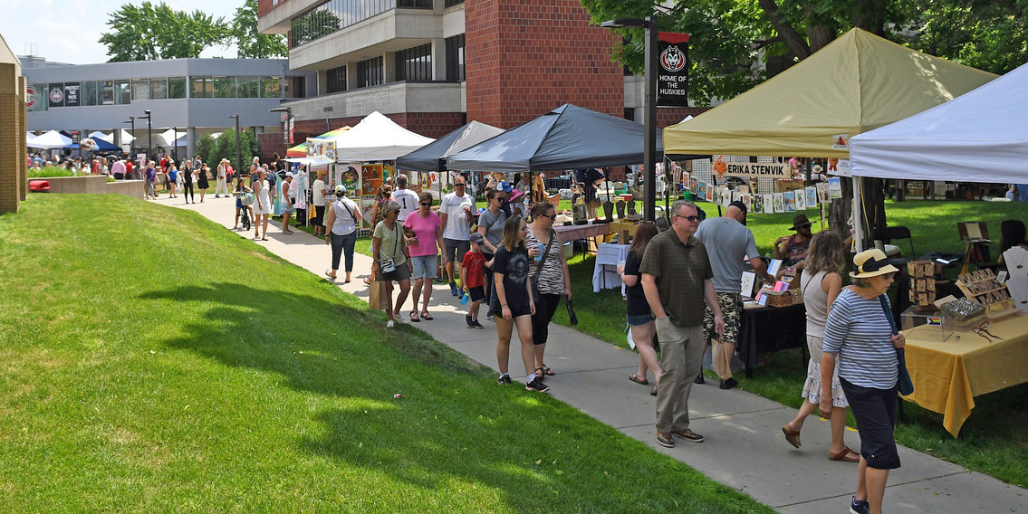 A crowd of people walks align a sidewalk with different crafts tables visible under tents. Atwood Memorial Center is visible in the background